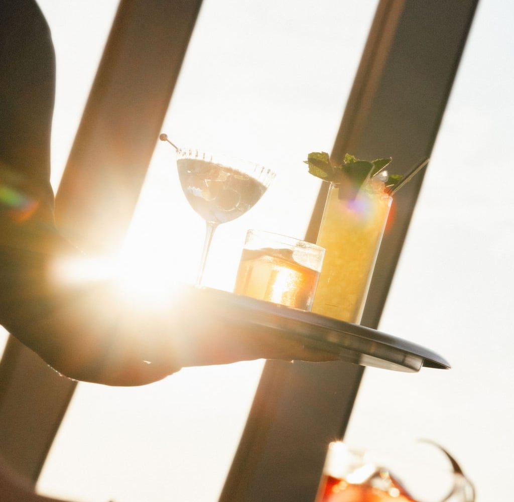 A server carries a tray of cocktails in front of bright sunset light, with a margarita and a tall garnished drink visible.