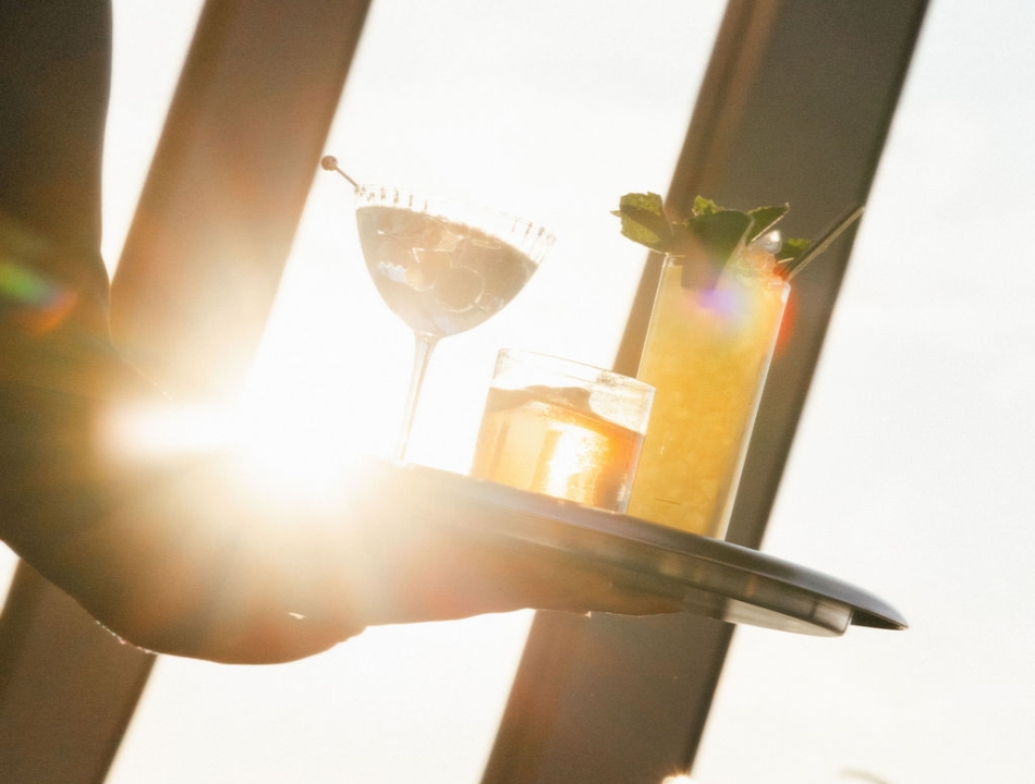 A server carries a tray of cocktails in front of bright sunset light, with a margarita and a tall garnished drink visible.