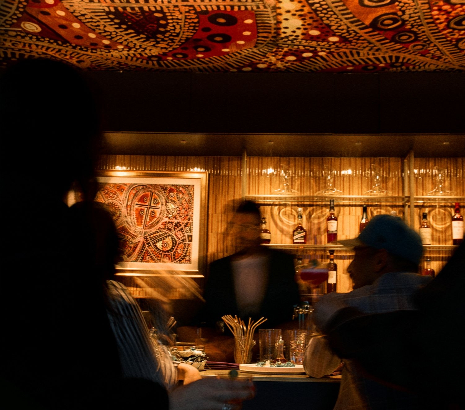Bar interior with warm lighting, blurred figures at a counter and bottles on backlit shelves in the background, decorative artwork on the wall above.