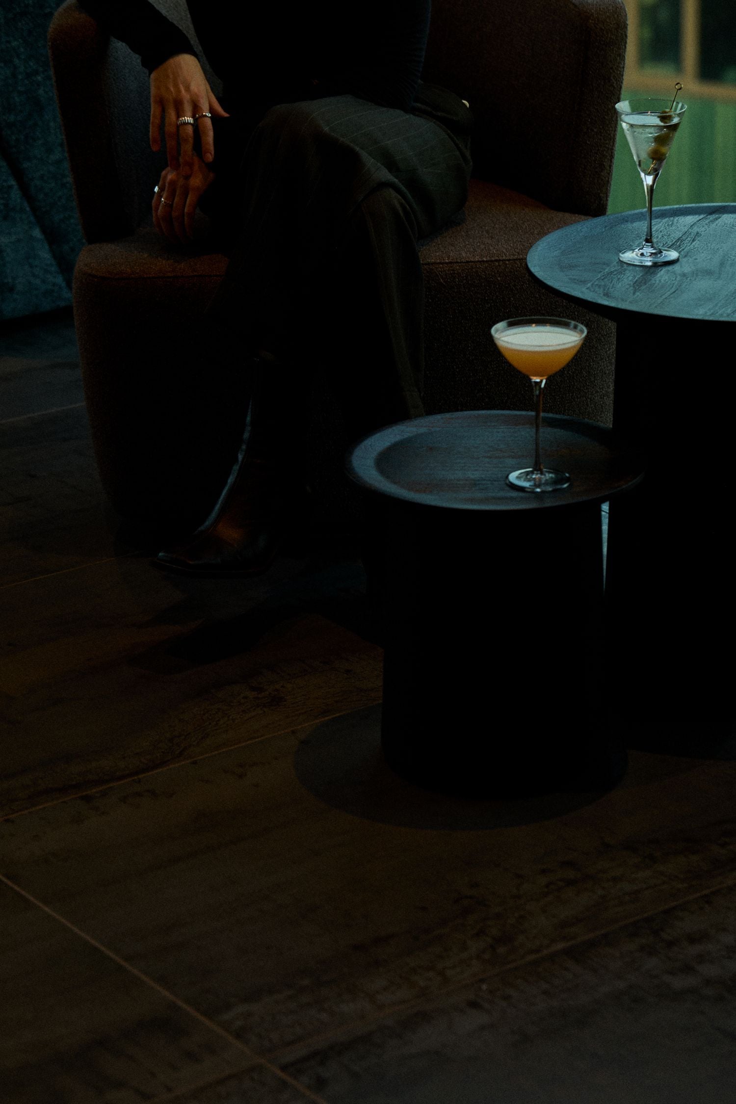 Dim, moody lounge scene with a person seated in dark clothing; hands with rings rest on the lap as two cocktails sit on round tables nearby.