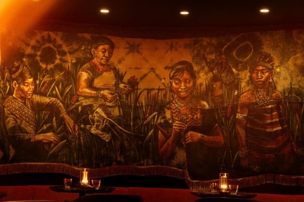 Dimly lit restaurant with a large mural of people in a field and candlelit dining tables, bathed in warm amber light.