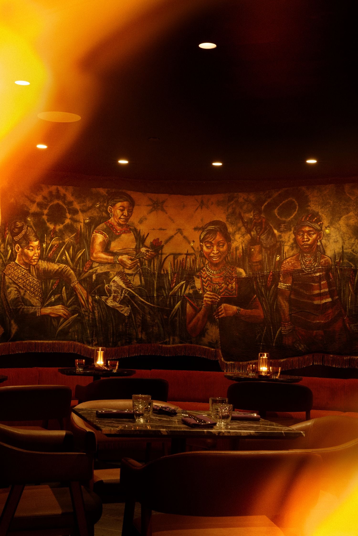 Dimly lit restaurant with a large mural of people in a field and candlelit dining tables, bathed in warm amber light.