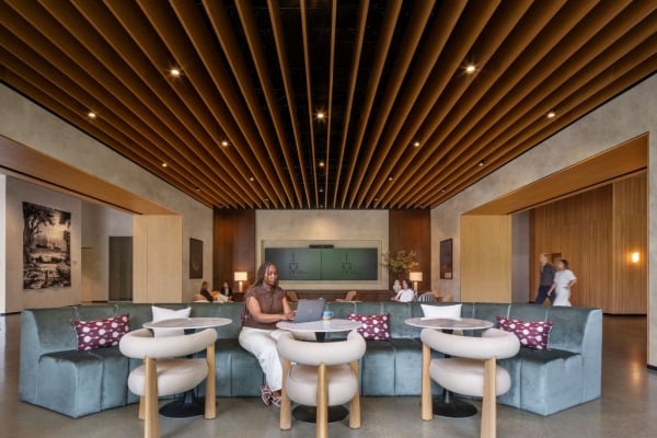 Person working on a laptop at a curved teal sofa lounge with round tables and a wooden slatted ceiling in a modern lobby.