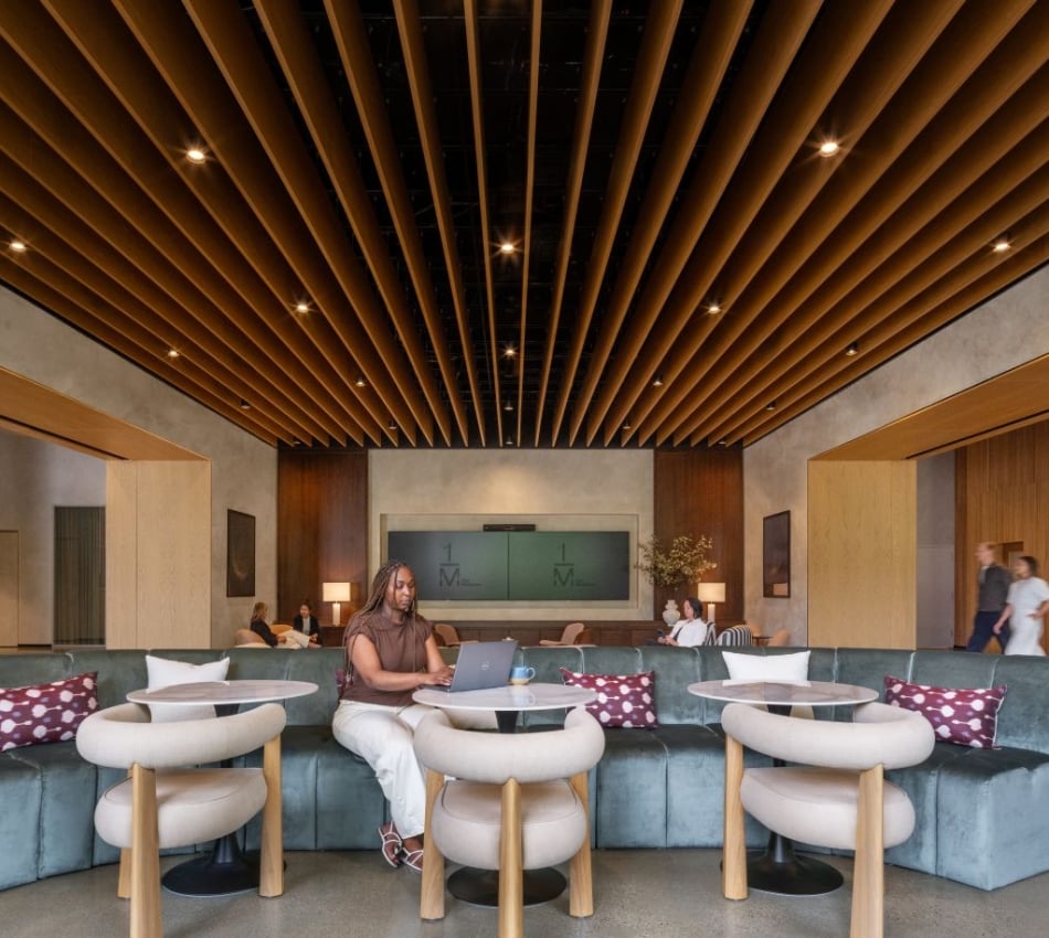 Person working on a laptop at a curved teal sofa lounge with round tables and a wooden slatted ceiling in a modern lobby.