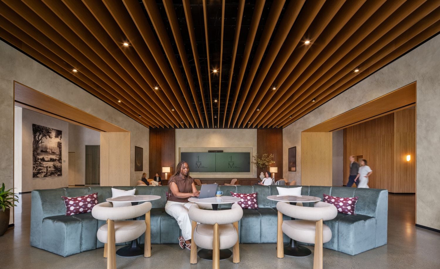 Person working on a laptop at a curved teal sofa lounge with round tables and a wooden slatted ceiling in a modern lobby.