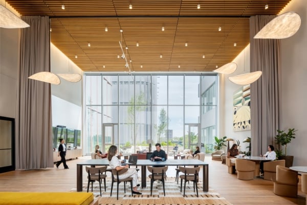 Open, airy hotel lobby or lounge with large glass walls, people seated at a long table and separate seating areas under a wooden slat ceiling.