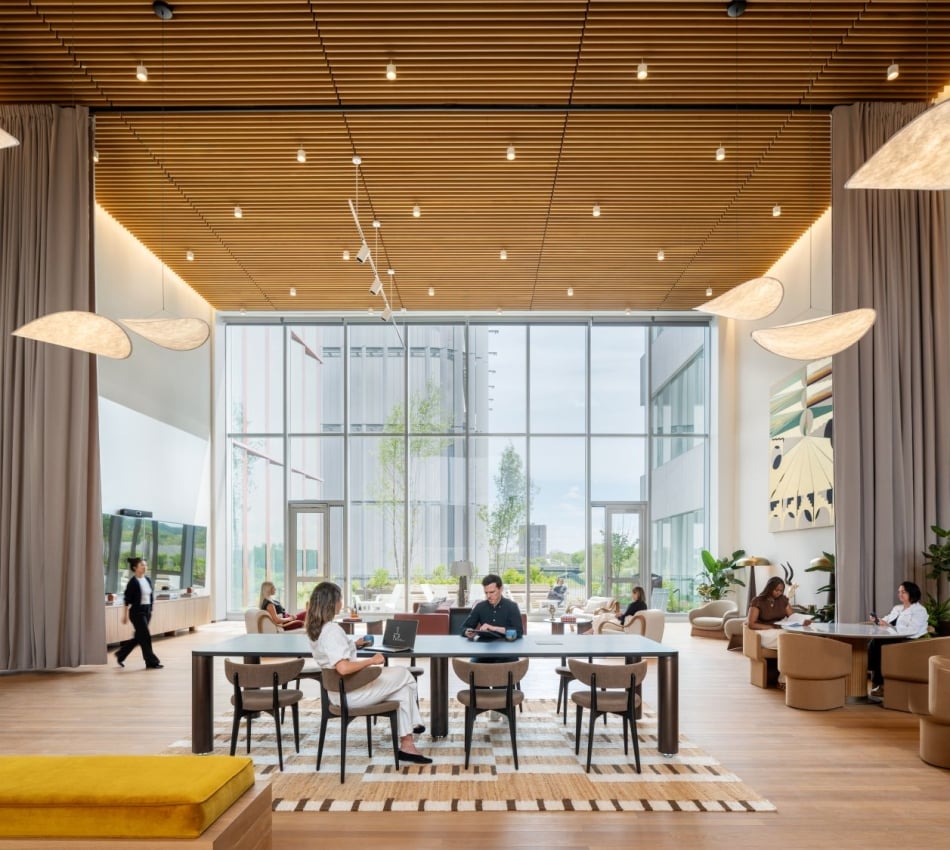 Open, airy hotel lobby or lounge with large glass walls, people seated at a long table and separate seating areas under a wooden slat ceiling.