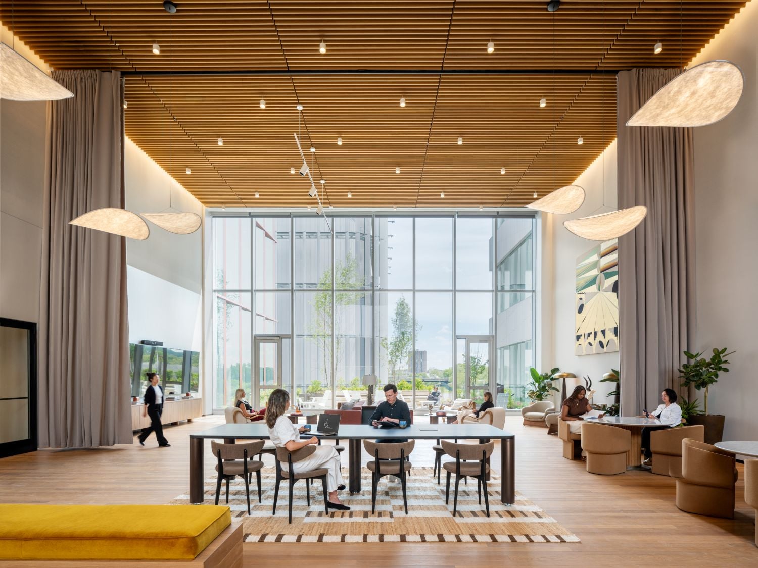 Open, airy hotel lobby or lounge with large glass walls, people seated at a long table and separate seating areas under a wooden slat ceiling.