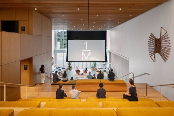 Modern lecture hall with a large front screen displaying 'One Milestone'; attendees sit on yellow upholstered benches facing the stage amid wooden walls and a slatted ceiling. A sunburst wall sculpture appears on the right.