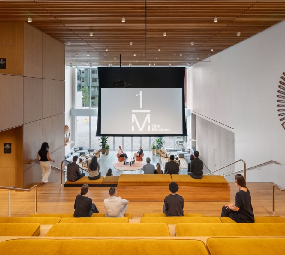 Modern lecture hall with a large front screen displaying 'One Milestone'; attendees sit on yellow upholstered benches facing the stage amid wooden walls and a slatted ceiling. A sunburst wall sculpture appears on the right.