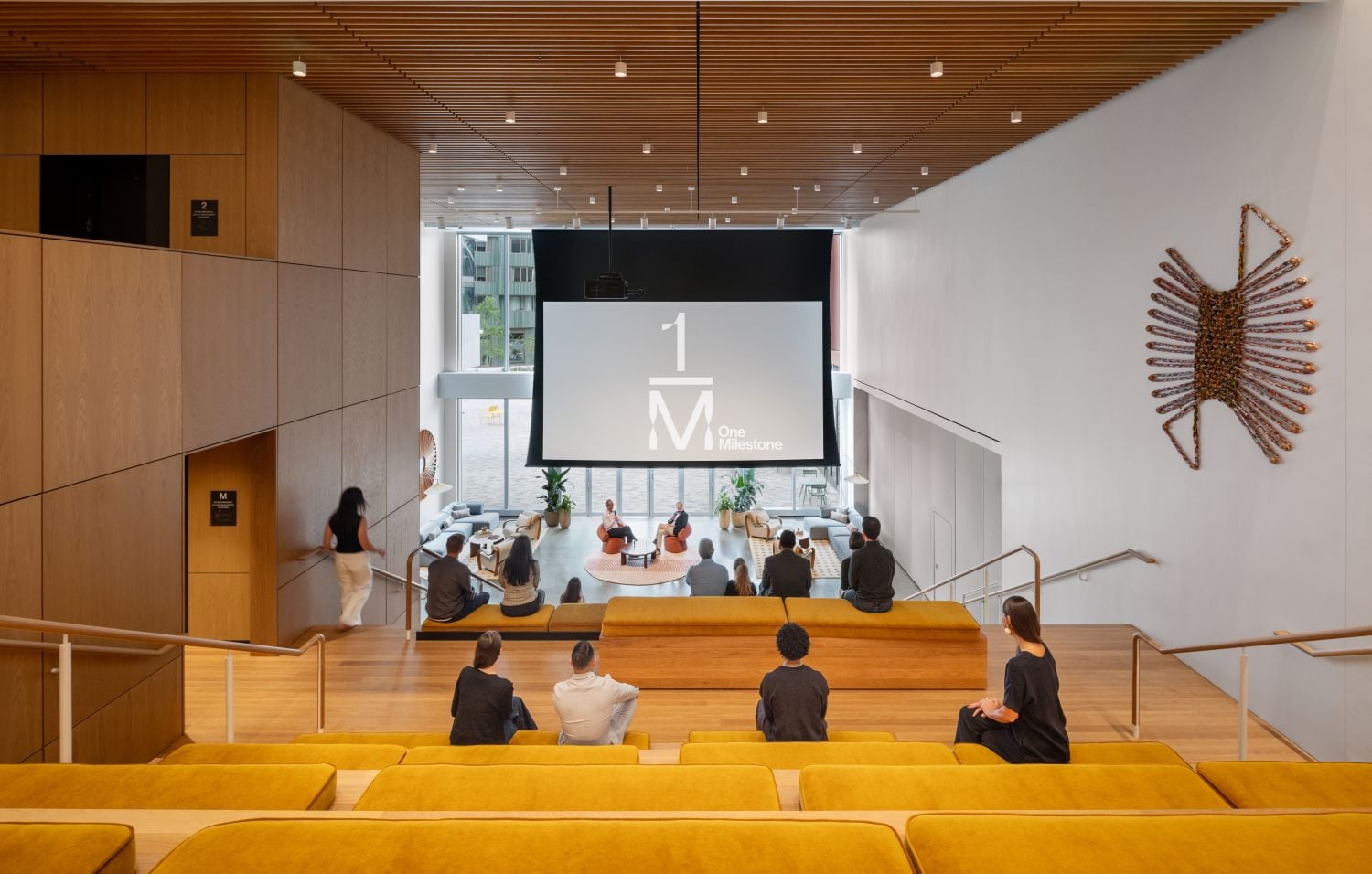 Modern lecture hall with a large front screen displaying 'One Milestone'; attendees sit on yellow upholstered benches facing the stage amid wooden walls and a slatted ceiling. A sunburst wall sculpture appears on the right.