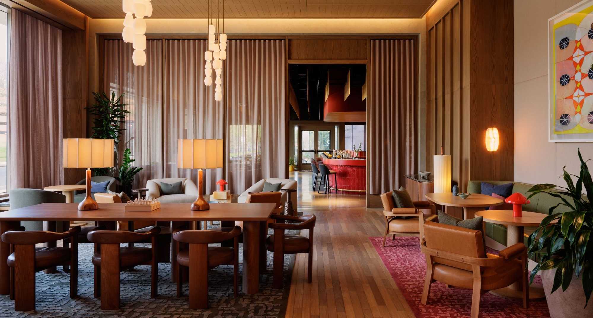 Spacious lounge with wooden tables and chairs, warm pendant lights, and tall curtains in a modern hotel-like lobby.