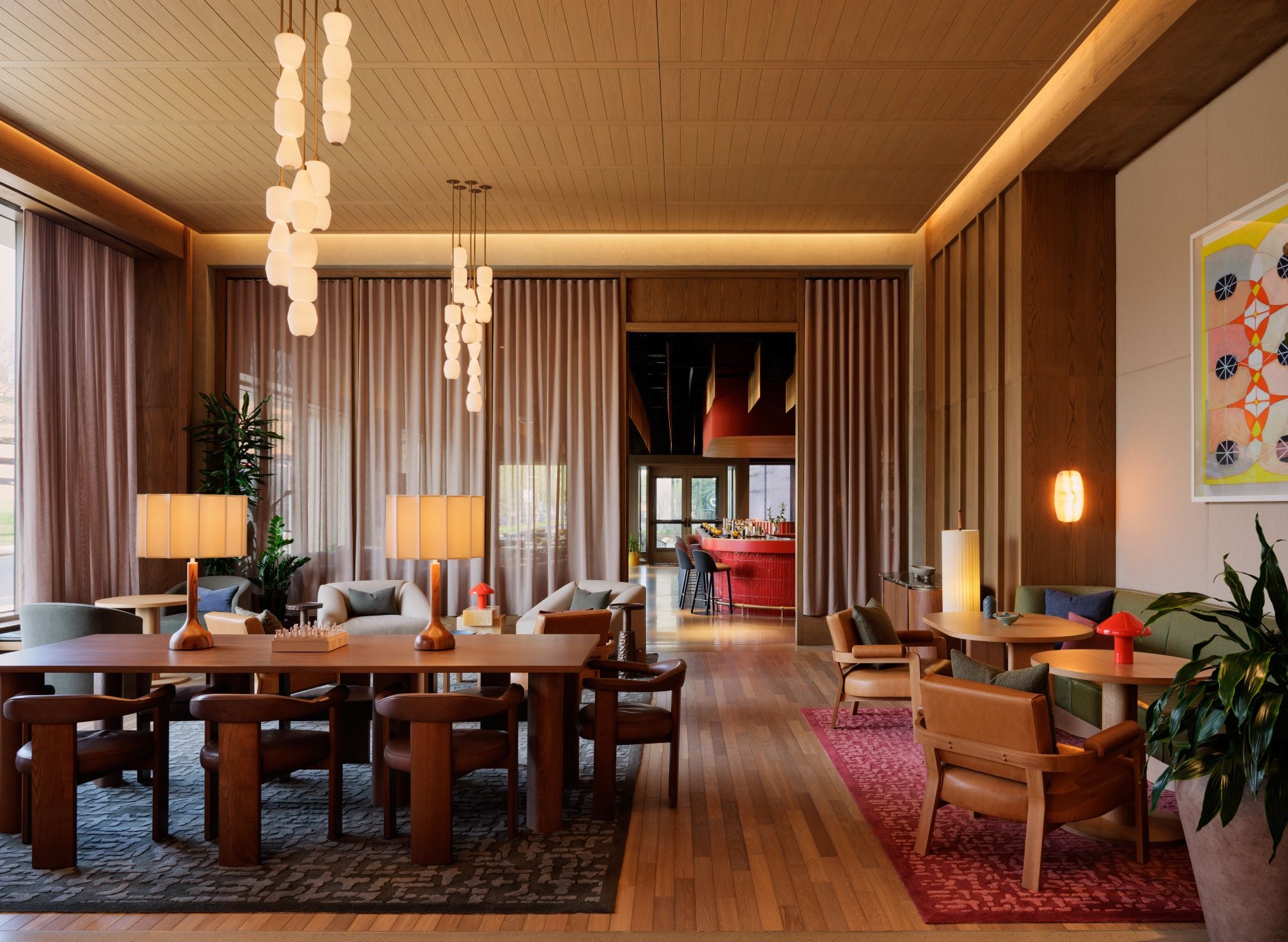 Spacious lounge with wooden tables and chairs, warm pendant lights, and tall curtains in a modern hotel-like lobby.