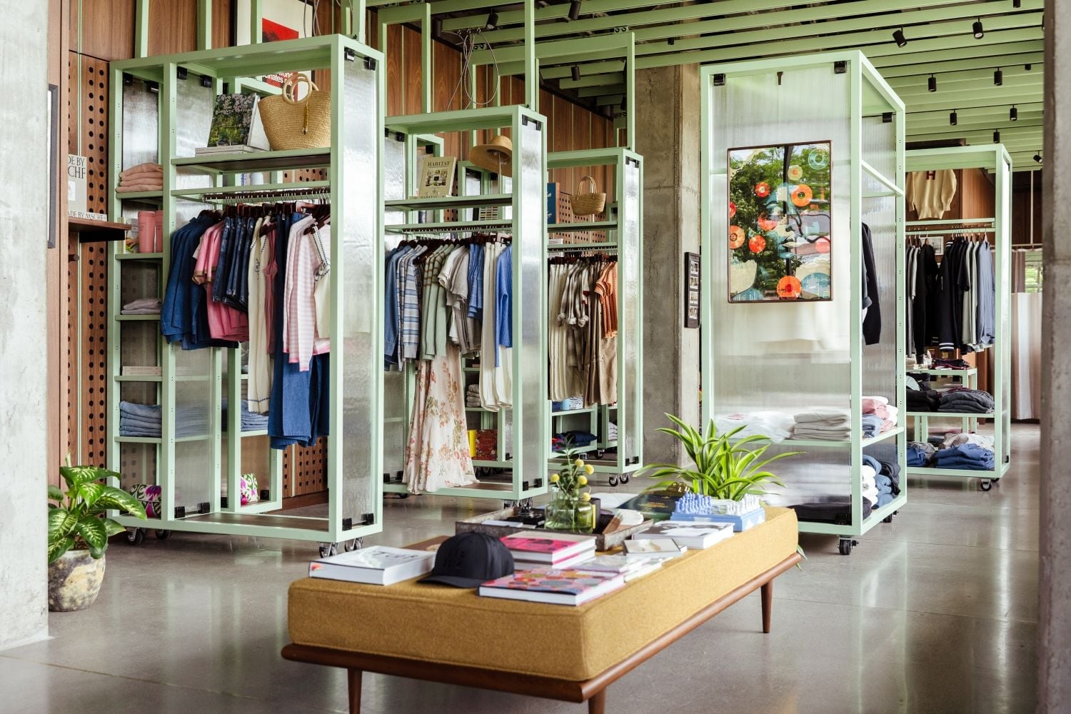 Mint-green clothing racks filled with shirts and dresses in a spacious boutique, with a beige magazine table in the foreground and plants around.