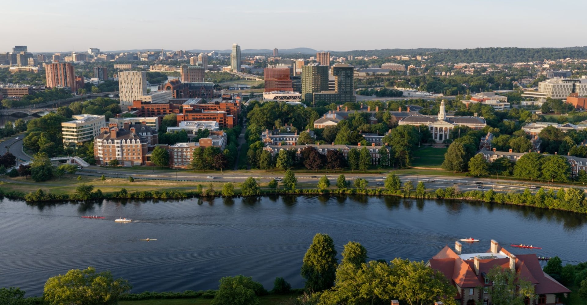 Aerial cityscape along a river with green trees, red brick and glass buildings, and boats on the water in the foreground.