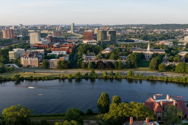 Aerial cityscape along a river with green trees, red brick and glass buildings, and boats on the water in the foreground.