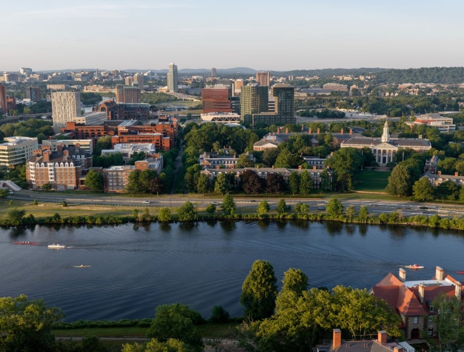 Aerial cityscape along a river with green trees, red brick and glass buildings, and boats on the water in the foreground.