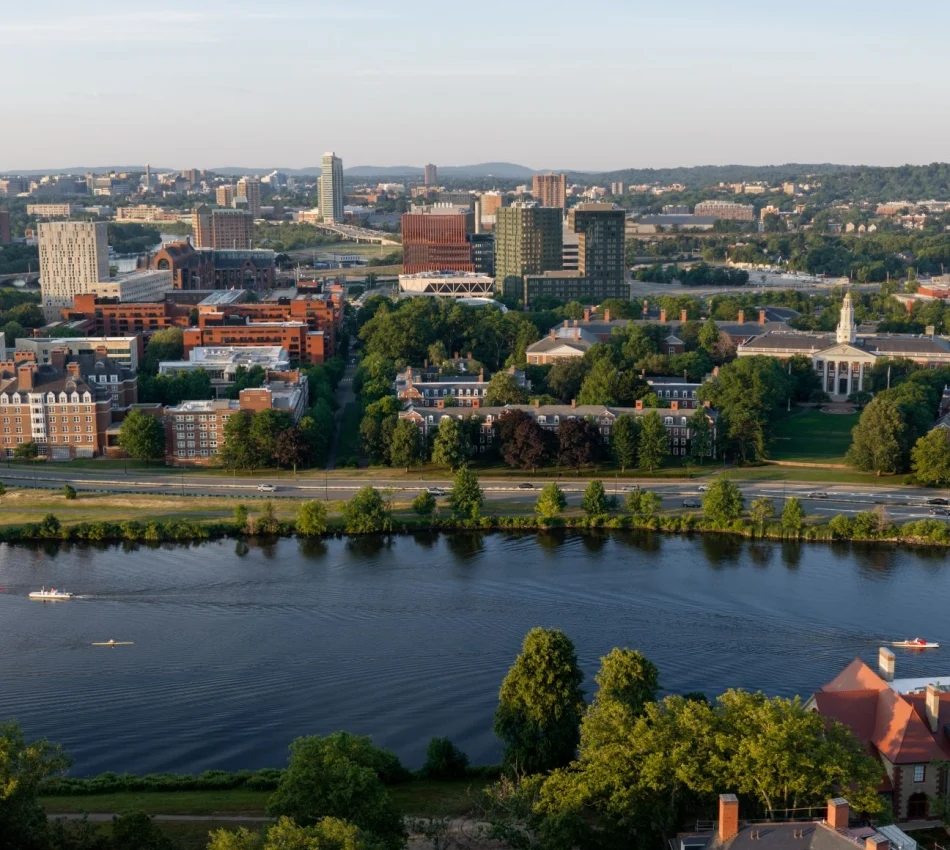 Aerial cityscape along a river with green trees, red brick and glass buildings, and boats on the water in the foreground.