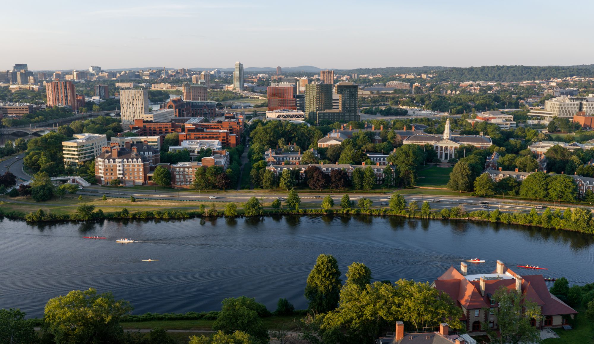 Aerial cityscape along a river with green trees, red brick and glass buildings, and boats on the water in the foreground.