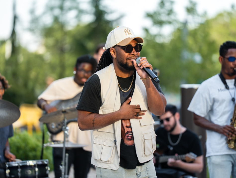 Male singer wearing a beige cap and vest, singing into a microphone on an outdoor stage with band members in the background.