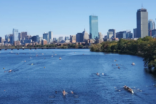 Boston skyline across the Charles River with rowers in small boats and a tree-lined riverbank under a clear blue sky