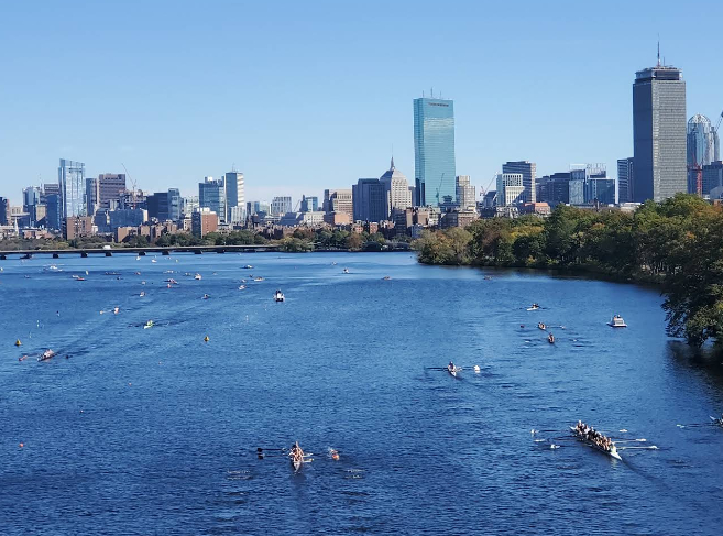 Boston skyline across the Charles River with rowers in small boats and a tree-lined riverbank under a clear blue sky
