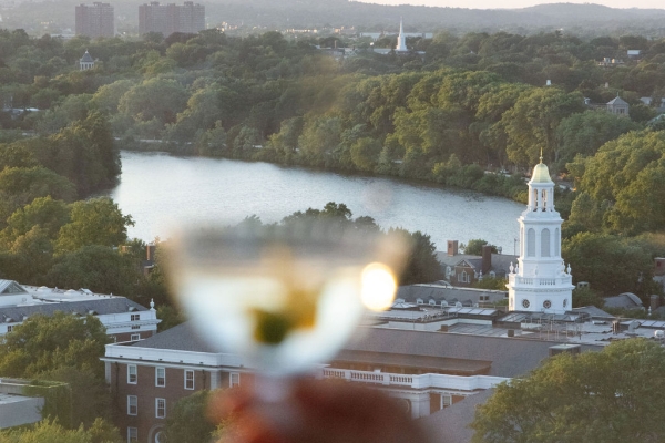 Blurred hand holding a glass of wine in the foreground, with a river, trees, and a white church steeple in a city skyline at sunset.