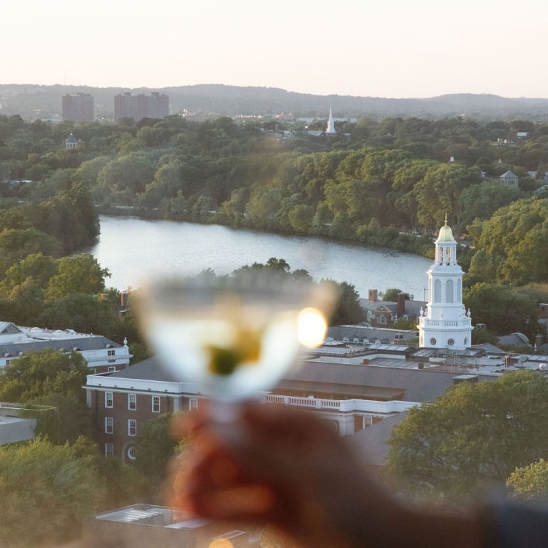 Blurred hand holding a glass of wine in the foreground, with a river, trees, and a white church steeple in a city skyline at sunset.