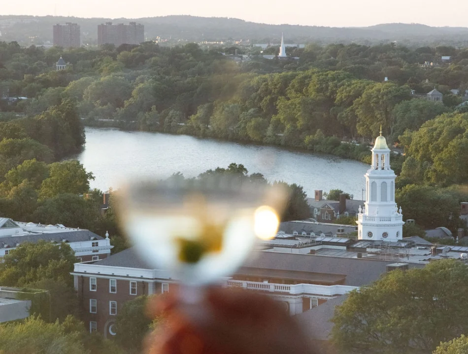 Blurred hand holding a glass of wine in the foreground, with a river, trees, and a white church steeple in a city skyline at sunset.