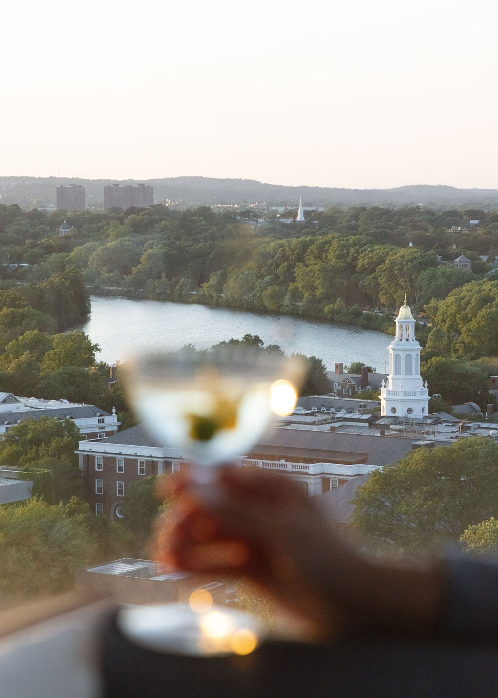 Blurred hand holding a glass of wine in the foreground, with a river, trees, and a white church steeple in a city skyline at sunset.