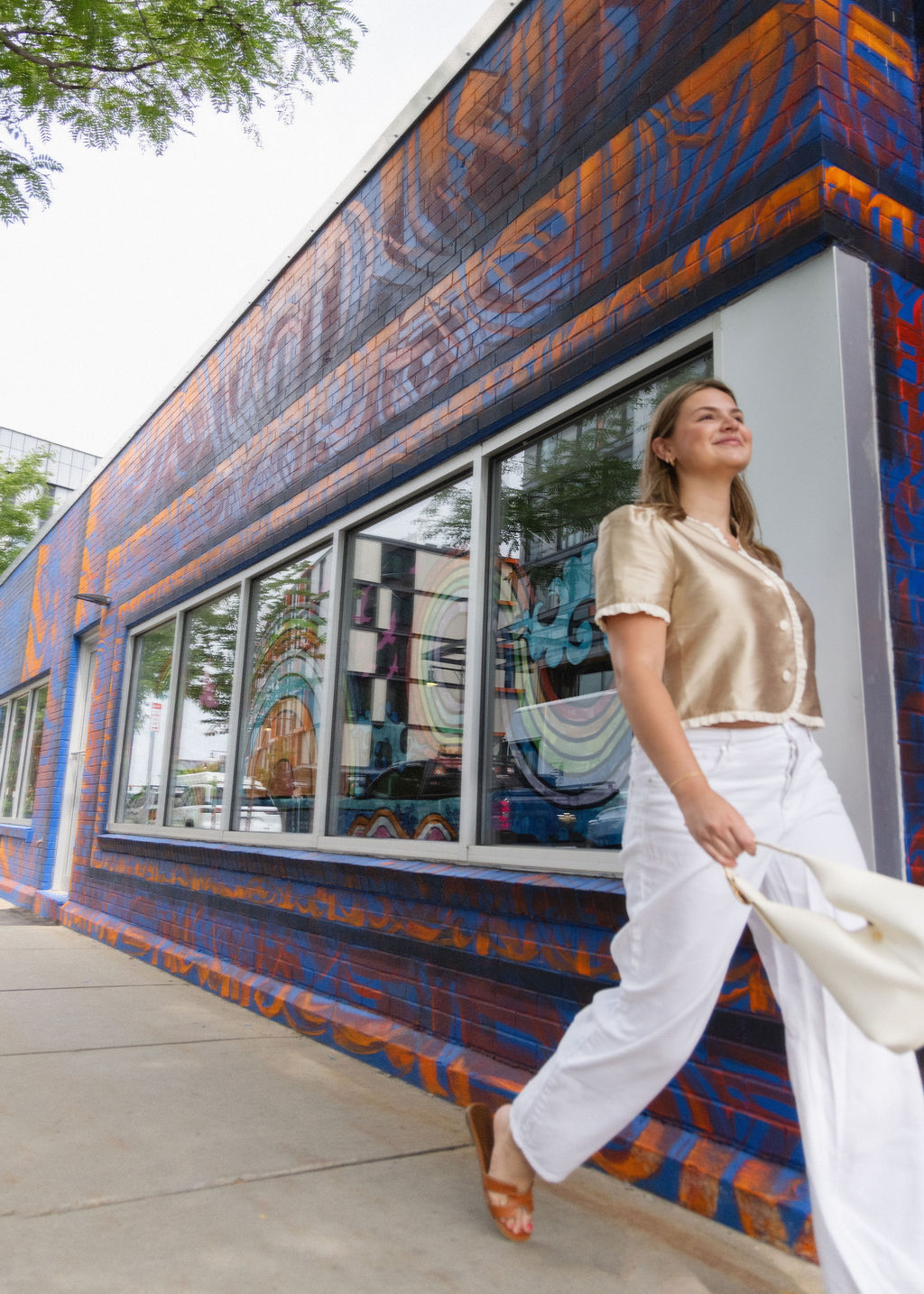 Woman walking past a colorful mural on a brick building, carrying a white bag, wearing a beige blouse and white pants.