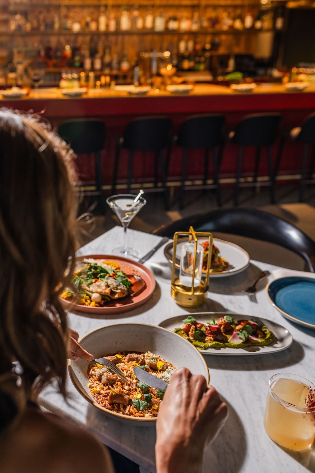 Diner at a marble table with three colorful plates and a cocktail, bar stools and bottles in the background.