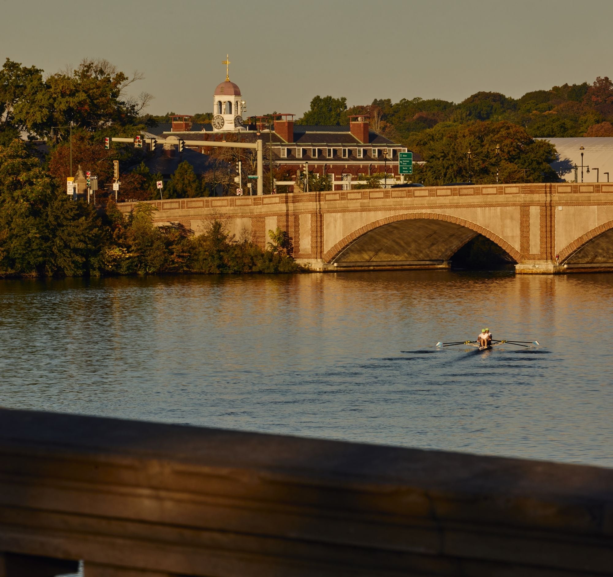 Two rowers in a small boat glide on a calm river as a brick arched bridge and brick building with a clock tower rise in the background at sunset.