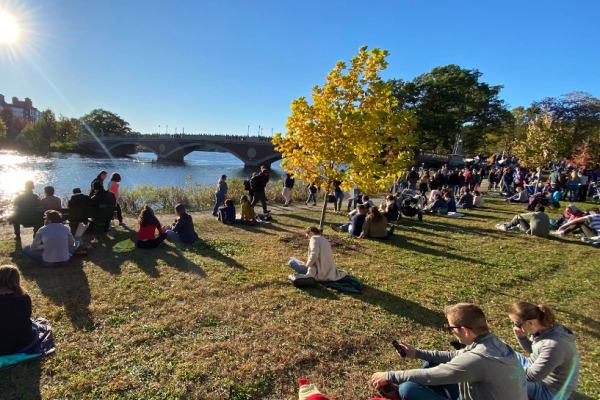 People picnicking and relaxing on a sunny riverside park with a stone bridge in the background and yellow autumn trees on the slope above the water.
