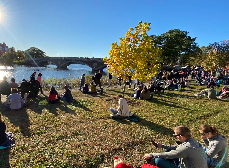 People picnicking and relaxing on a sunny riverside park with a stone bridge in the background and yellow autumn trees on the slope above the water.