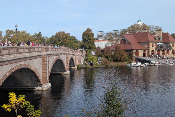 Crowded brick arch bridge over a calm river, with a red-roofed riverside building and boats docked at the shore.