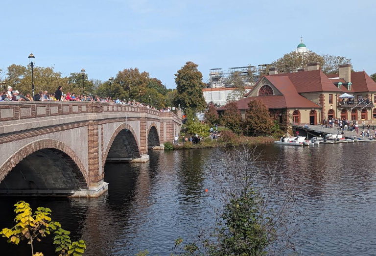 Crowded brick arch bridge over a calm river, with a red-roofed riverside building and boats docked at the shore.
