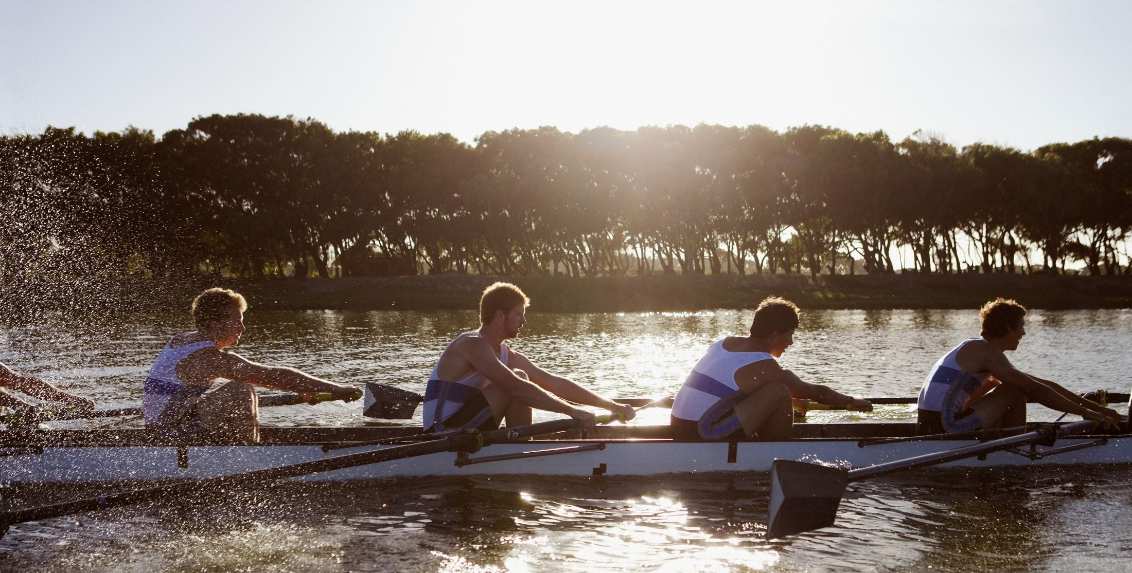 Four rowers in a racing shell glide across a sunlit lake with a tree-lined shore in the background, splashing water.