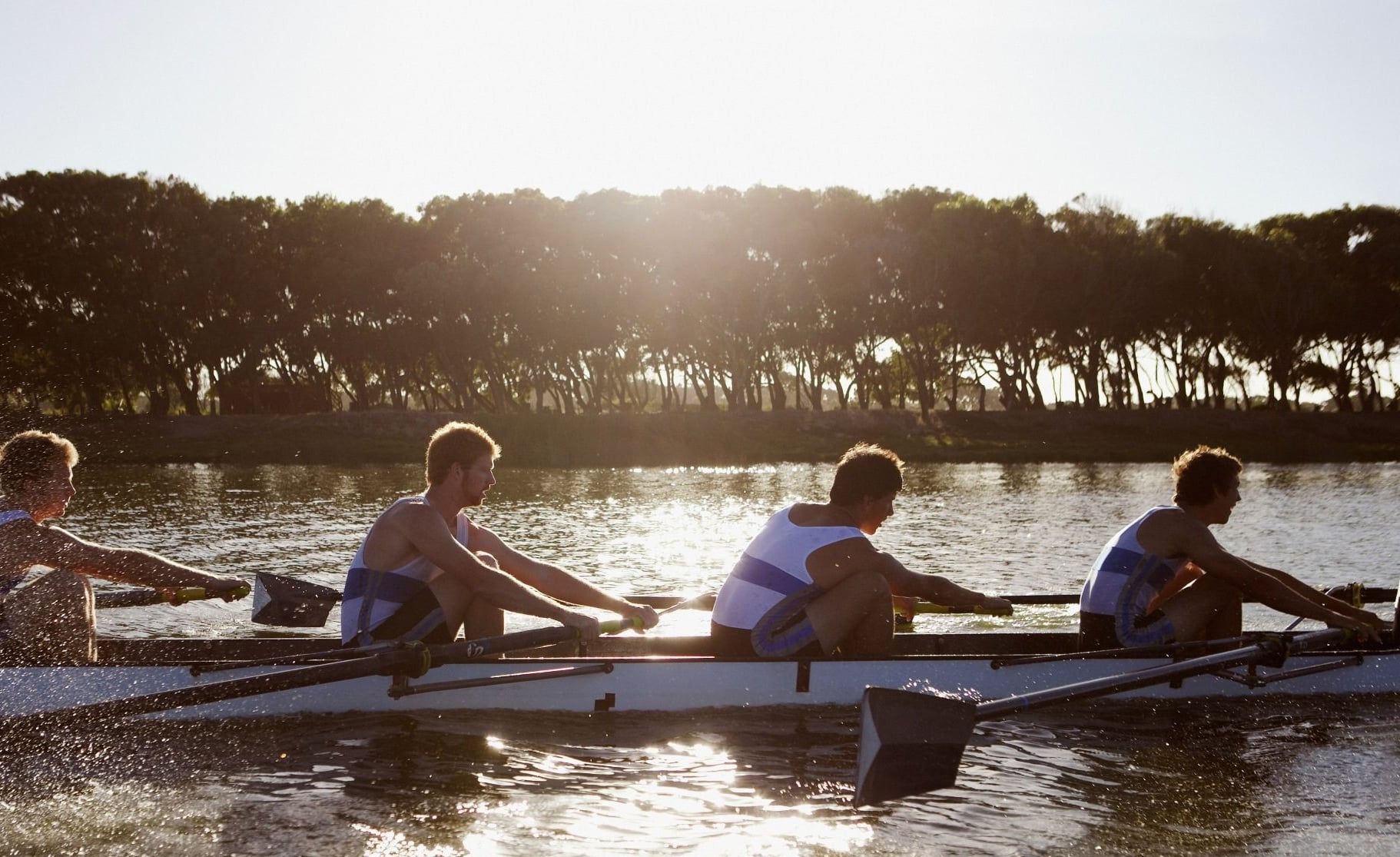 Four rowers in a racing shell glide across a sunlit lake with a tree-lined shore in the background, splashing water.