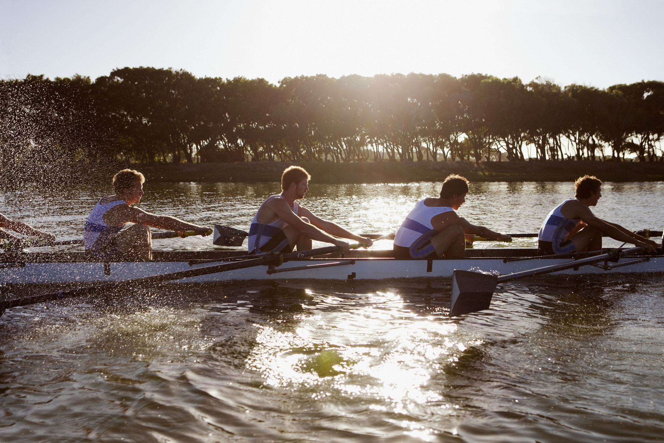 Four rowers in a racing shell glide across a sunlit lake with a tree-lined shore in the background, splashing water.
