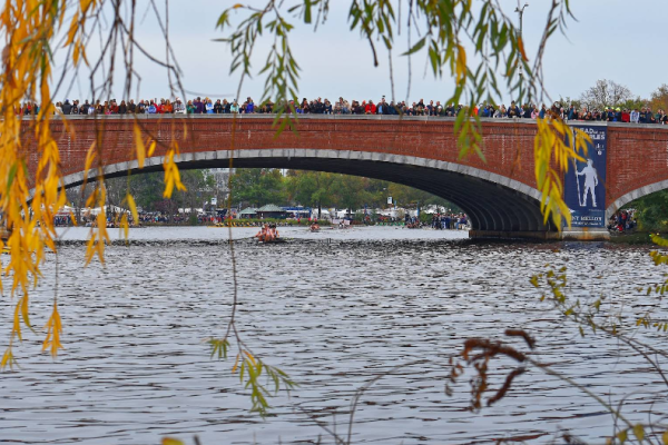 Crowded brick arch bridge over a river during an outdoor event, framed by autumn leaves in the foreground.
