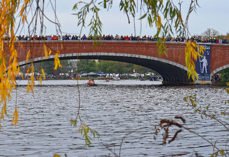 Crowded brick arch bridge over a river during an outdoor event, framed by autumn leaves in the foreground.