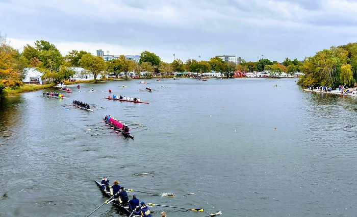 Overhead view of a rowing regatta on a wide river; several eight-oared shells in competition with colorful uniforms, spectators along the riverbank, and trees showing autumn colors in the background.