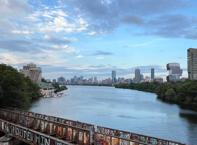 Graffiti-covered bridge railing in the foreground, calm river, and a distant city skyline under a blue sky.