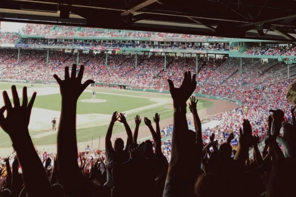 Crowd of baseball fans with hands raised in the foreground at a daytime game in a crowded stadium.