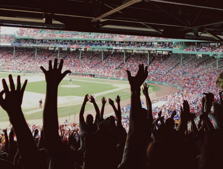 Crowd of baseball fans with hands raised in the foreground at a daytime game in a crowded stadium.