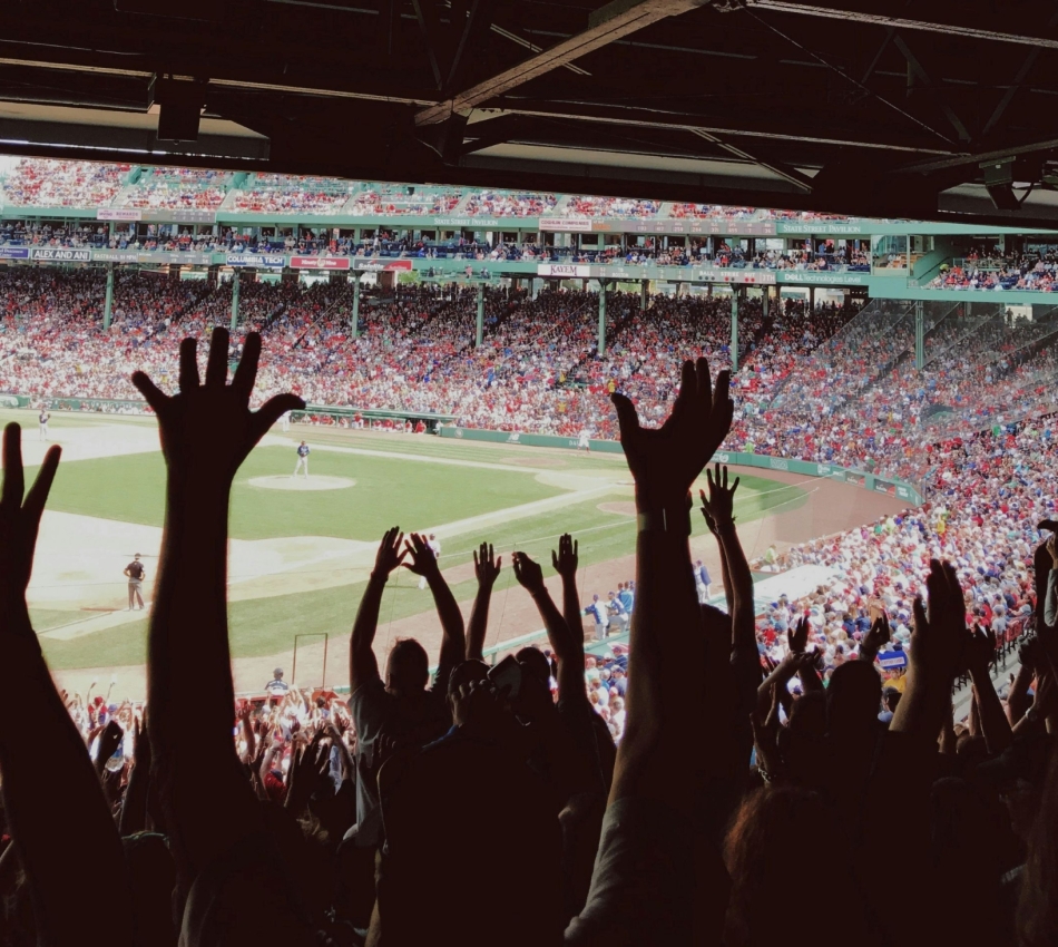Crowd of baseball fans with hands raised in the foreground at a daytime game in a crowded stadium.