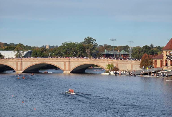 Stone arch bridge over a wide river with many people on the walkway and rowers on the water.