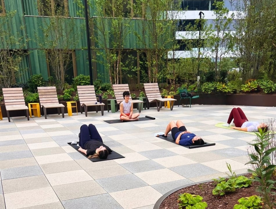 Outdoor yoga class: four participants on mats in a tiled courtyard with an instructor seated cross‑legged in the center, greenery and modern building in the background.
