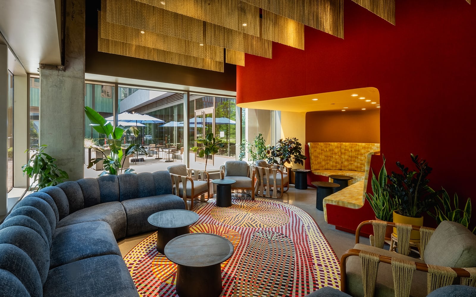 A modern hotel lounge with a curved blue sofa, colorful patterned rug, and seating areas by large glass walls overlooking outdoor tables with umbrellas.